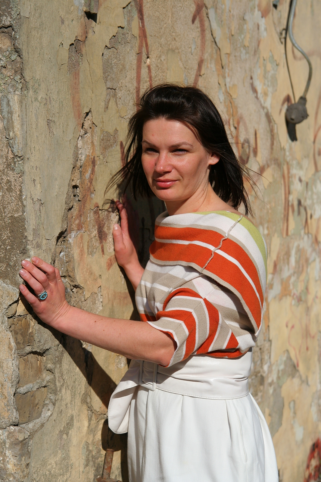 A woman with straight, dark hair poses against a weathered, textured wall with peeling paint. She wears an orange and white striped top and a white skirt, her left hand resting on the wall. Gazing directly at the camera in bright, natural sunlight, she embodies a modern mode talka vibe.