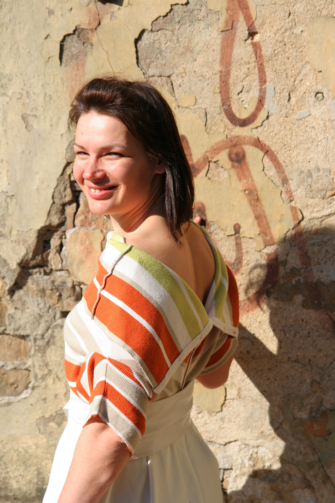 A smiling woman stands against a weathered wall with peeling paint and faded graffiti. She has shoulder-length dark hair and wears an off-the-shoulder striped top in orange, green, and white colors, paired with a white skirt. The sunlight casts subtle shadows, highlighting her cheerful expression as she enjoys her modes talka surroundings.