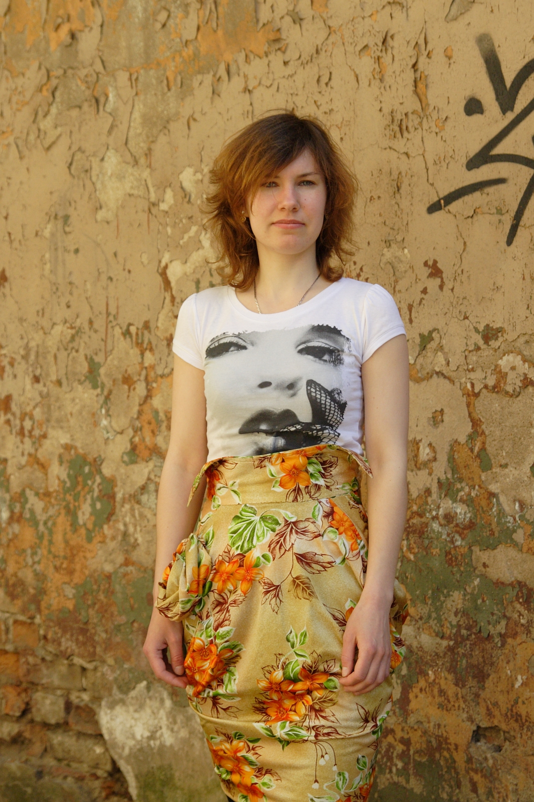 A person with shoulder-length hair stands against a worn, graffiti-marked wall. They wear a white T-shirt featuring a face with a butterfly and a patterned skirt with orange flowers and leaves. The wall, evocative of various artistic modes, has peeling paint revealing patches of brick and plaster.