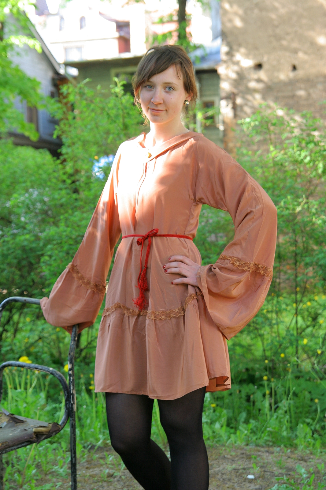 A woman with short brown hair stands outside in front of greenery and a building. She is wearing a loose-fitting peach-colored dress with long, billowy sleeves, cinched at the waist with a red cord. With one hand on her hip and the other resting on the back of a metal chair, she exudes modes talka.