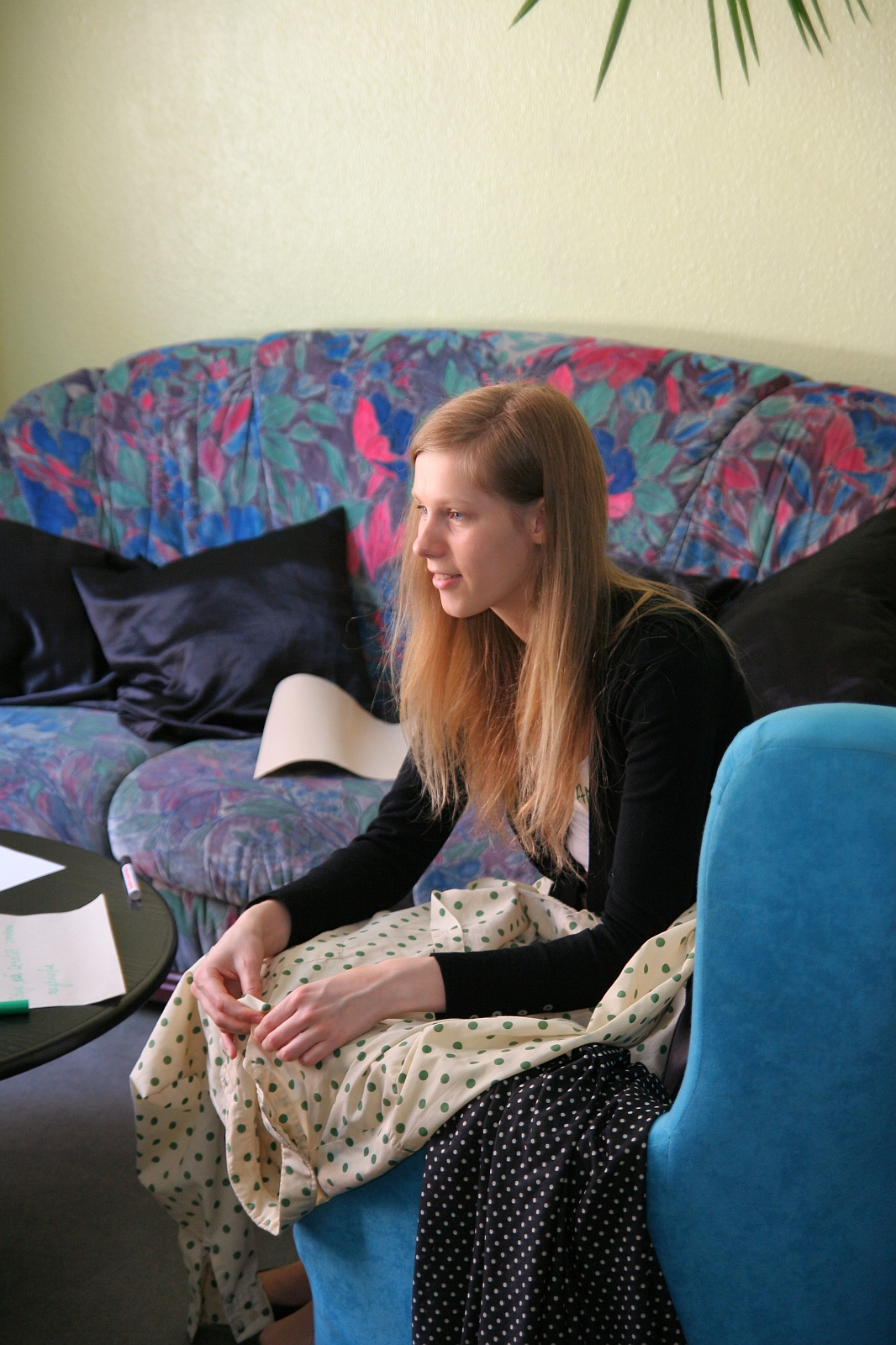 A woman with long blonde hair is sitting on a blue chair, wearing a black top and a white polka-dotted skirt. She is smiling and looking to her right. In the living room, with its colorful floral-patterned couch and black cushions behind her, sits a table scattered with papers from her latest Modes talka.