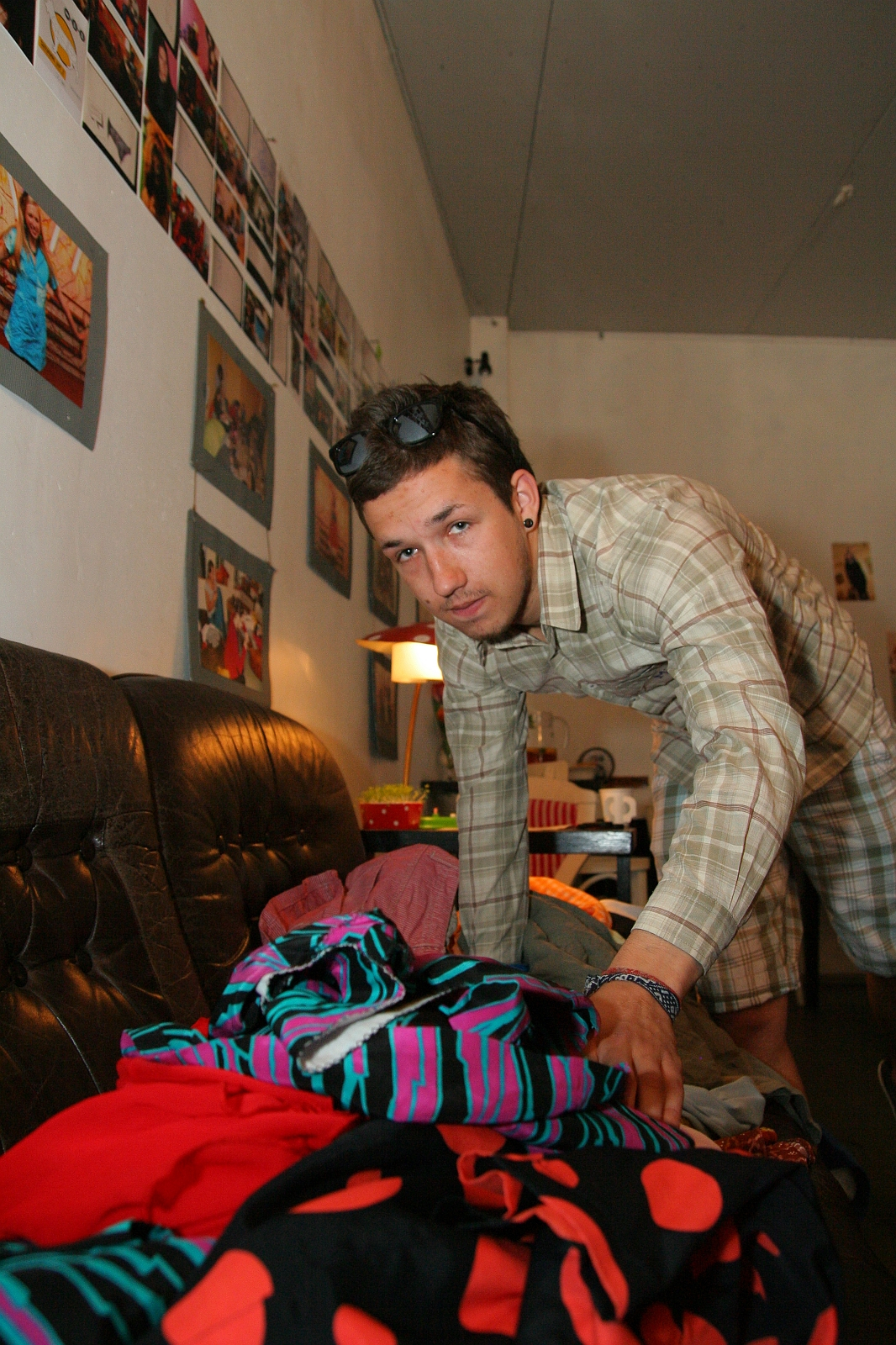 A person wearing a plaid shirt and plaid shorts leans over a sofa, sorting through a pile of colorful clothes. The room is decorated with various photographs and posters on the walls. There is a lamp on a small table behind the person, its light showcasing their modes talka collection.