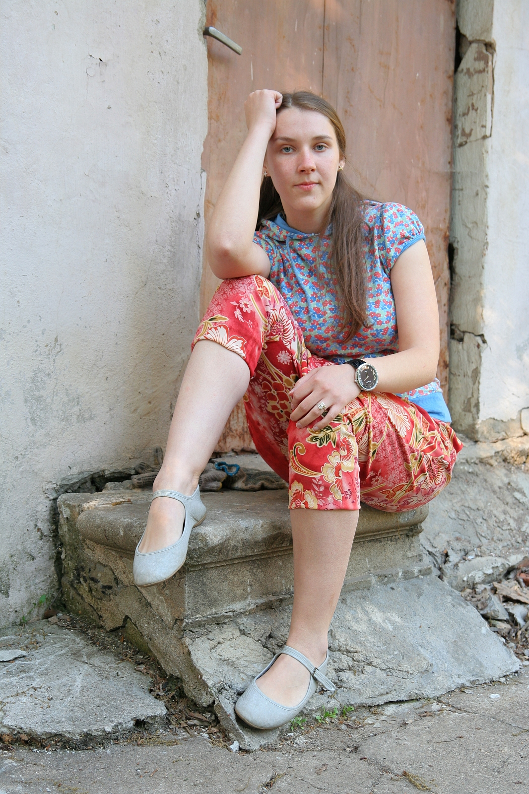 A young woman with long, brown hair, wearing a blue floral shirt and red floral pants, sits in a talka mood on a small concrete step against a weathered wall and door. She rests her left elbow on her knee and her head on her hand, looking into the camera. She wears gray flats and a wristwatch.