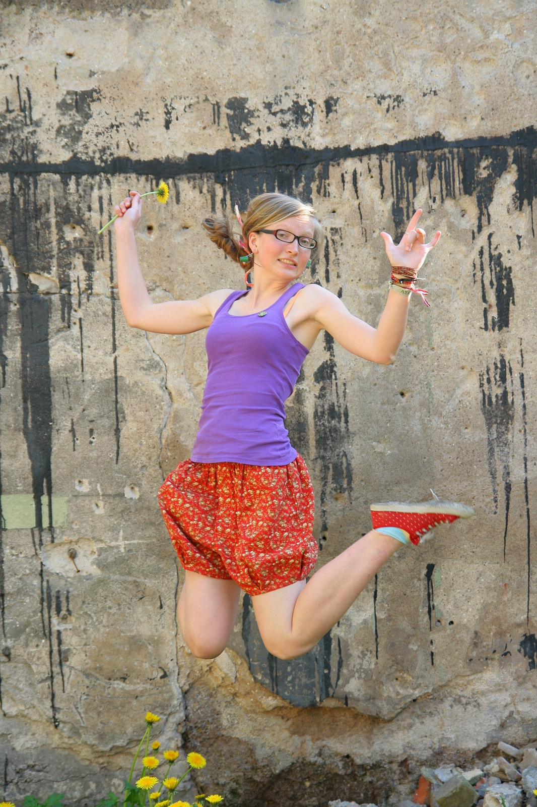 A woman with glasses, wearing a purple tank top and red floral skirt, jumps joyfully against a grungy, textured wall with black paint streaks. She holds a small flower in one hand and wears multiple bracelets. Her hair is in a side ponytail, and her red shoes with white polka dots add flair to the scene.