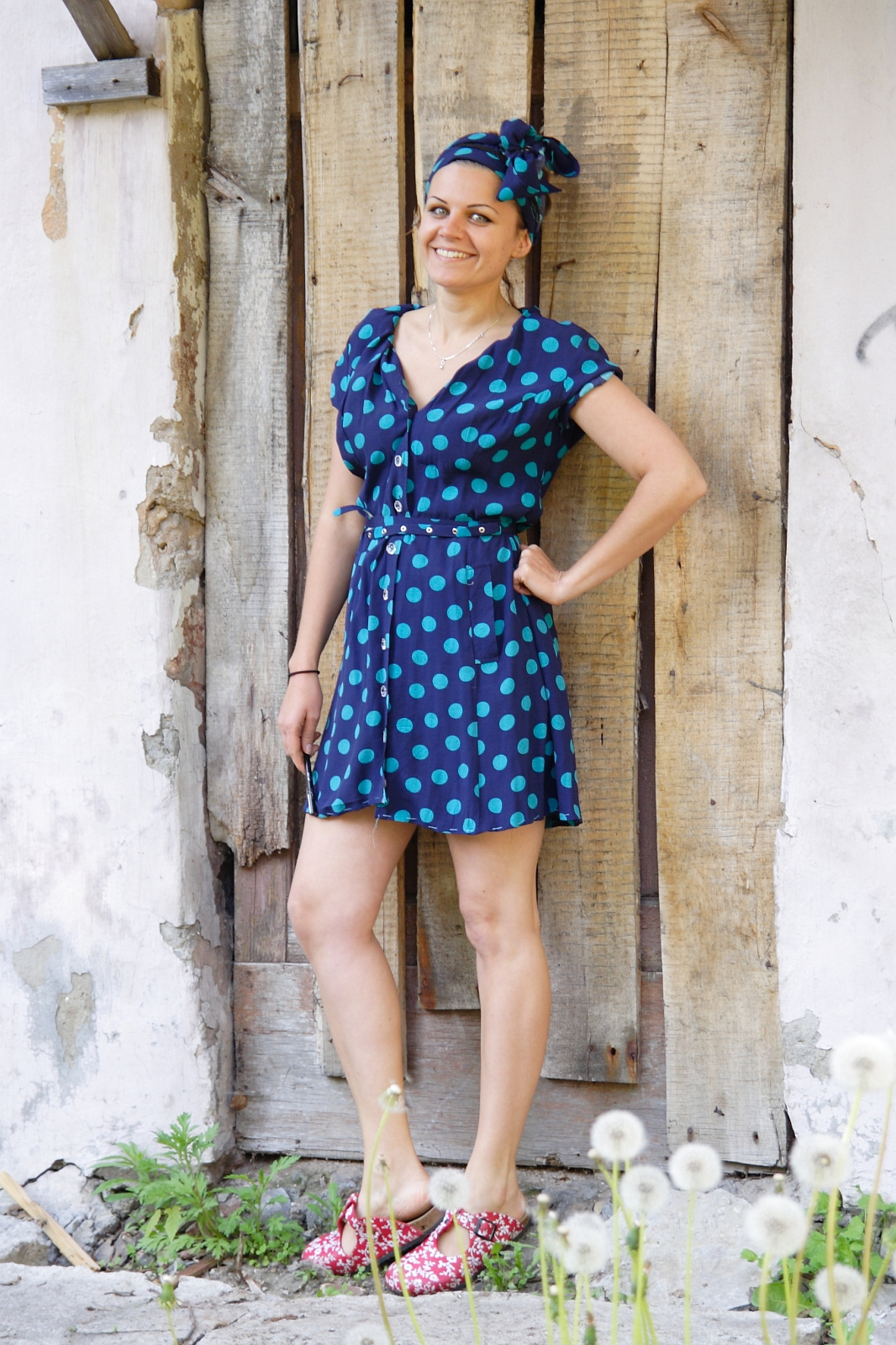 A smiling woman stands in front of a rustic wooden door, engaging in a bit of modes talka with passersby. She wears a blue dress with green polka dots, accessorized with a matching headscarf. Her left hand is on her hip, and she has on red patterned sandals. The weathered wall and dandelions in the foreground add to the picturesque setting.