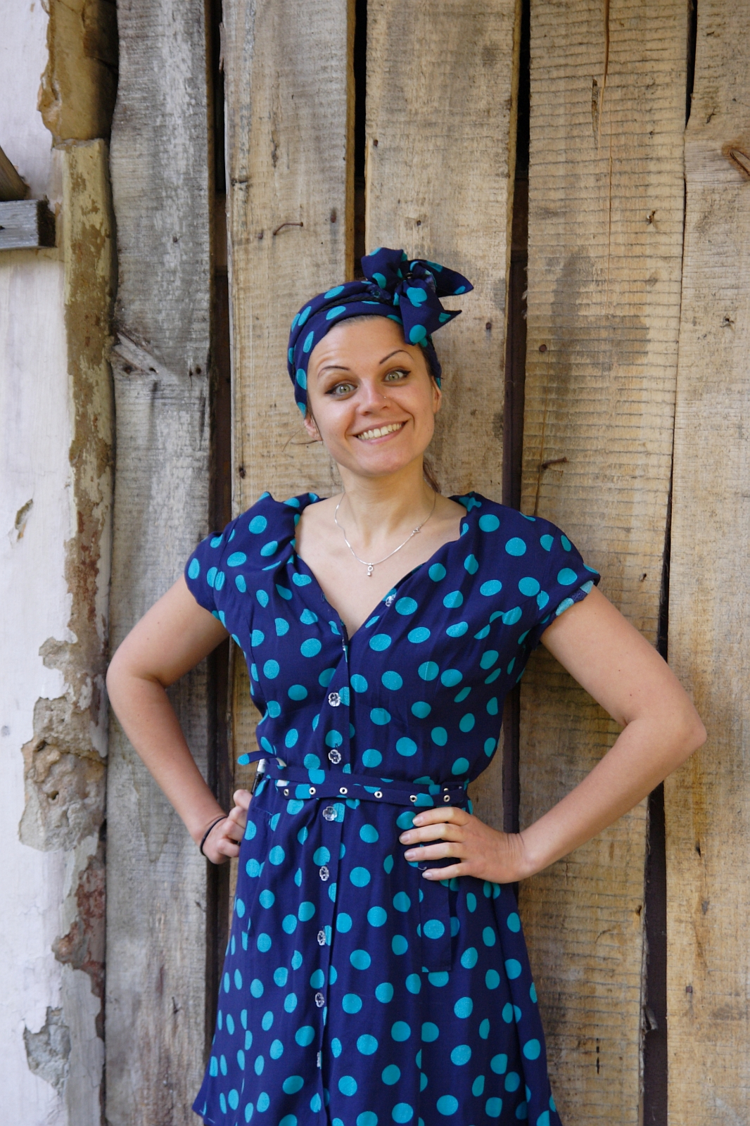 A person stands smiling with hands on hips. They are wearing a blue dress with large turquoise polka dots, a matching belt, and a headscarf in the same pattern. The background features a rustic wooden wall with varying shades and textures, adding charm to their stylish mode of dress.