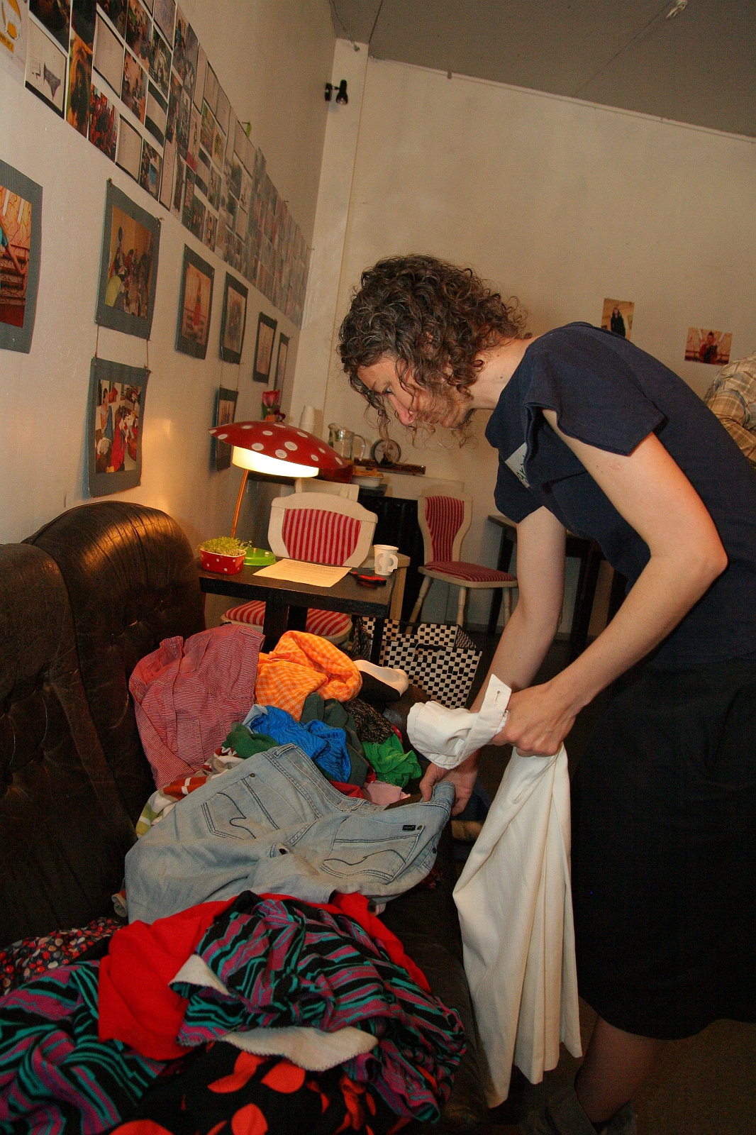 A person with curly hair, wearing a dark blue dress, is sorting through a pile of colorful clothes on a brown sofa. The room has a white wall adorned with framed photos and modes under a red lamp. Behind them, a table with red and white chairs and talka miscellaneous items are visible.