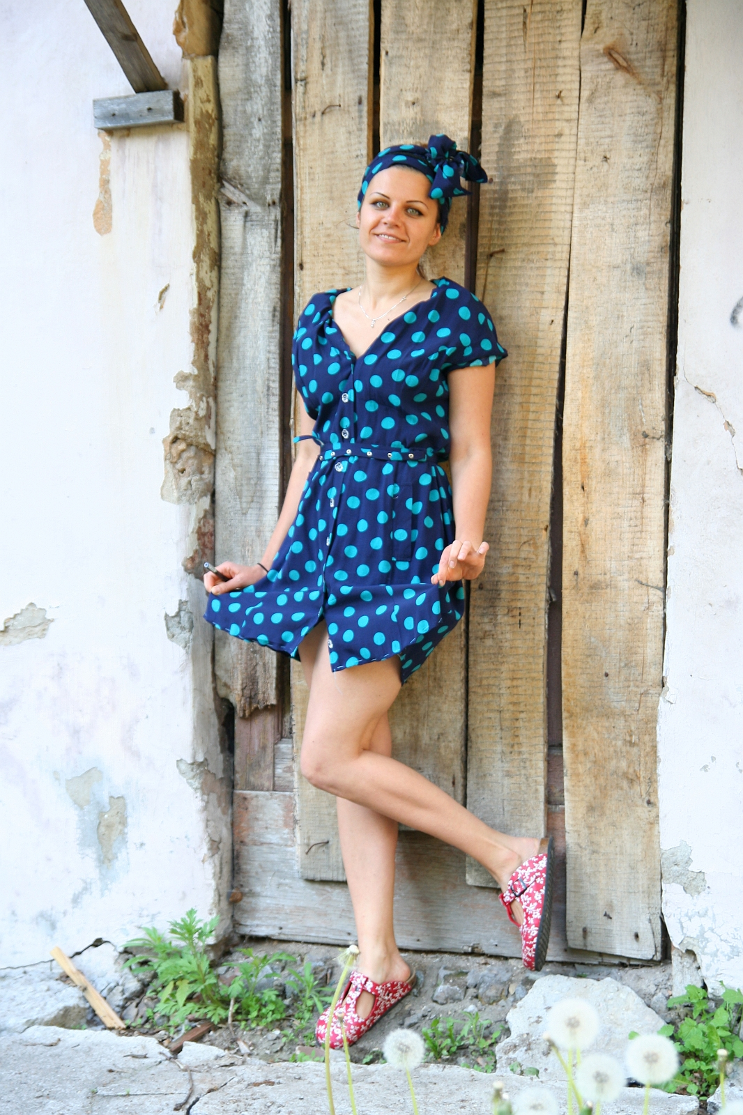 A woman stands against a rustic wooden door, wearing a blue dress with green polka dots and a matching headscarf by Modes Talka. She has her left foot raised, displaying floral-patterned shoes. Her hands are playfully tugging at her dress as she smiles warmly. The background features weathered wood and stone.