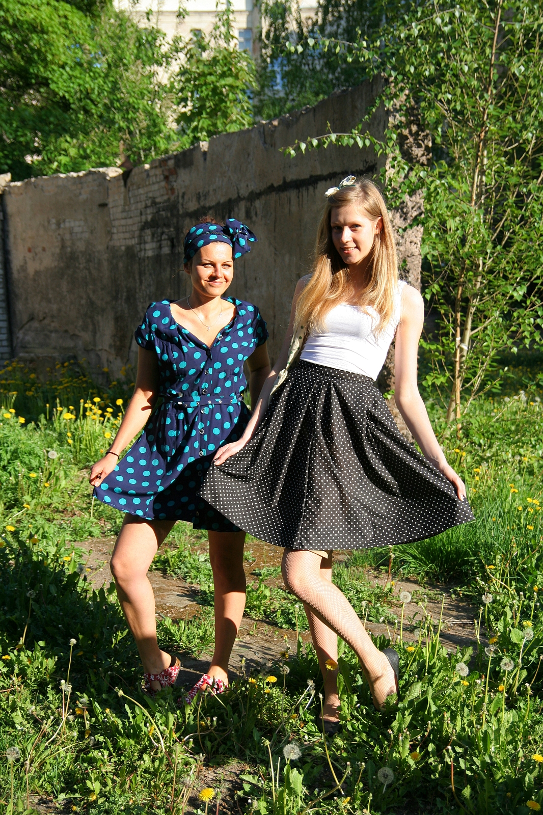 Two young women stand in a sunlit, overgrown garden. The woman on the left wears a blue polka-dot dress and headscarf, smiling. The woman on the right wears a white tank top and black polka-dot skirt, also smiling. Both hold onto their skirts, creating an airy, playful atmosphere amidst the green talka.