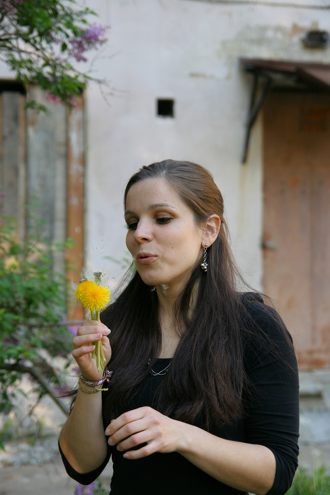 A woman with long brown hair wearing a black top is standing outdoors and blowing on a dandelion. The background shows an old building with a wooden door and greenery, including a flowering tree to the left. Her expression appears focused on the modes talka as the dandelion seeds disperse into the air.