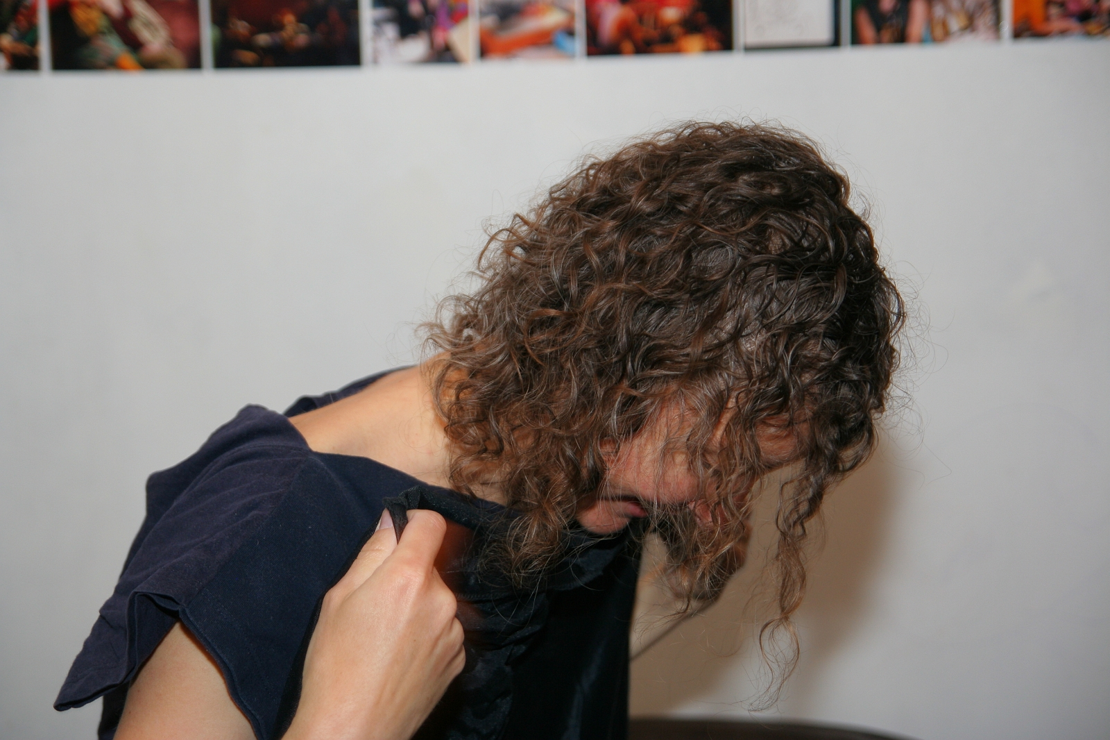 A person with curly brown hair is partially lifting a dark shirt, covering part of their face, and looking downward. The background shows a white wall adorned with various small, colorful photographs along the upper edge—a perfect backdrop for a modes talka vibe.