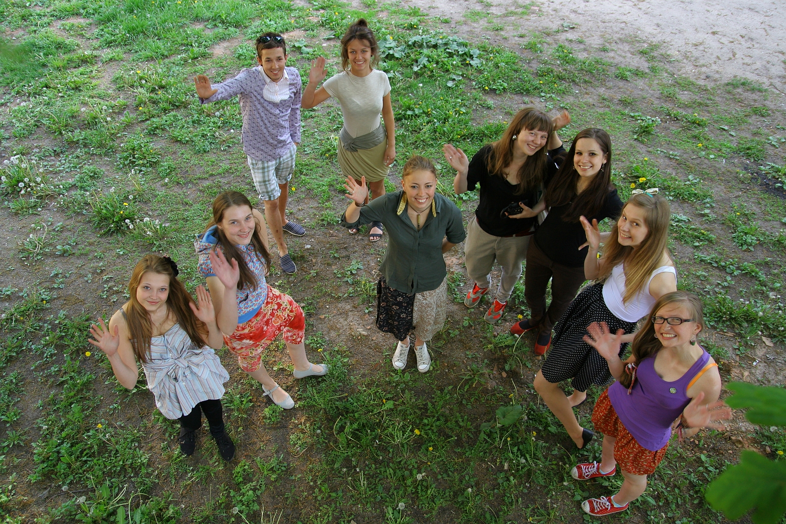 A group of eight people, consisting of six women and two men, is standing in a semi-circle on a grassy area outdoors, engaged in lively Modes talka. They are all facing the camera and waving, smiling brightly. The background shows a mix of green grass and patches of dirt. The atmosphere appears joyous and casual.