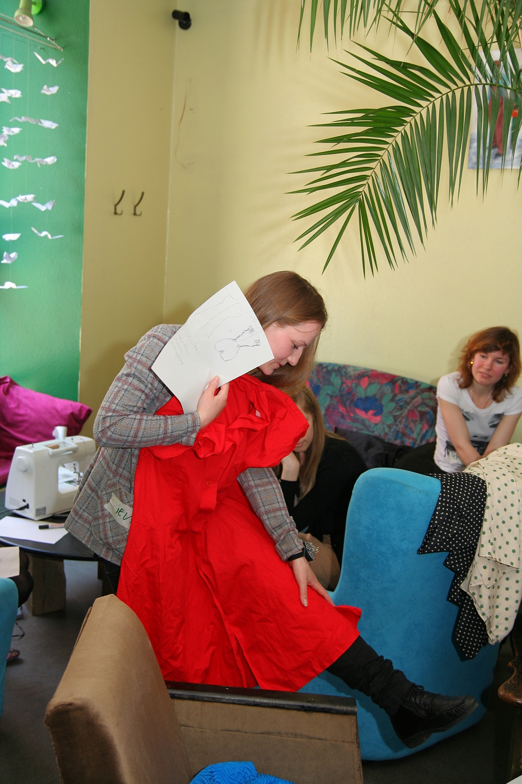A person holding a paper and a red garment while sitting on a blue chair in a room with green walls and various fabrics and decorations. Other individuals are present, one on another blue chair. A sewing machine is visible on a table in the background, along with Modes patterns scattered around. A large plant is partly visible.