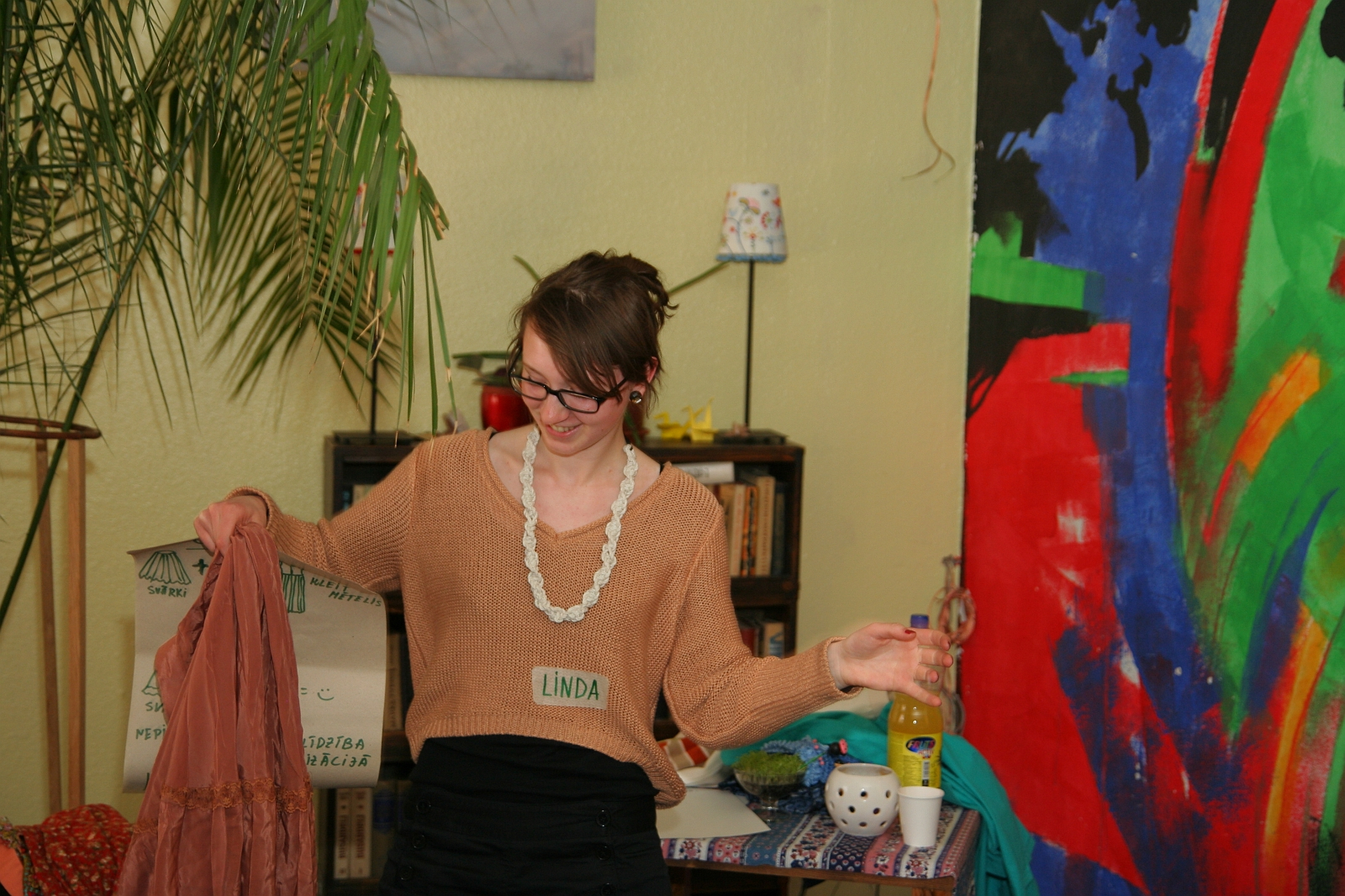 A person wearing a light brown sweater with a "Linda" name tag, white beaded necklace, and eyeglasses holds a peach-colored cloth in one hand and a clear bottle with yellow liquid in the other. They stand in Modes Talka, surrounded by vibrant abstract painting, bookshelves, and various decorations.