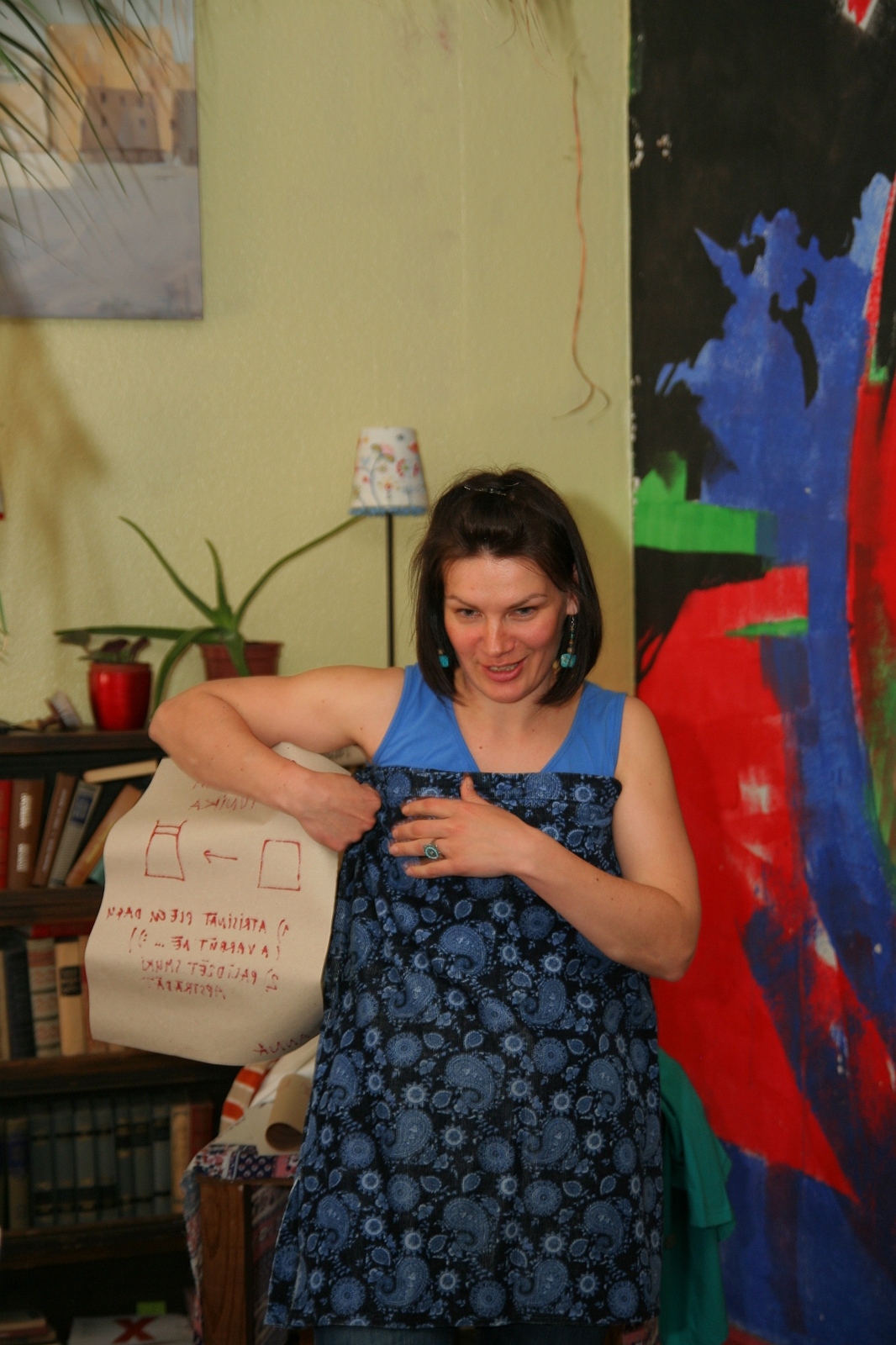 A woman with short dark hair, wearing a blue patterned dress and turquoise earrings, is standing indoors near a bookshelf and a large colorful wall painting. Sporting a cloth bag with a sketch and "Modes talka" text on it, she partially covers her outfit as various houseplants adorn the space.