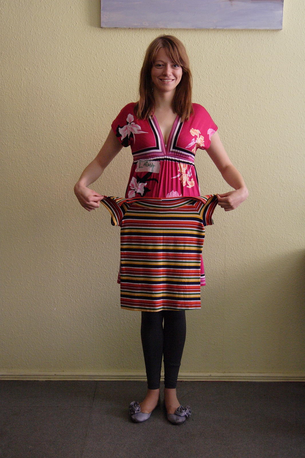 A woman with shoulder-length hair, wearing a pink floral dress over black leggings and grey flats, is standing indoors against a beige wall. She is smiling and facing the camera while holding up a colorful striped dress, showcasing her vibrant choice in modes talka.