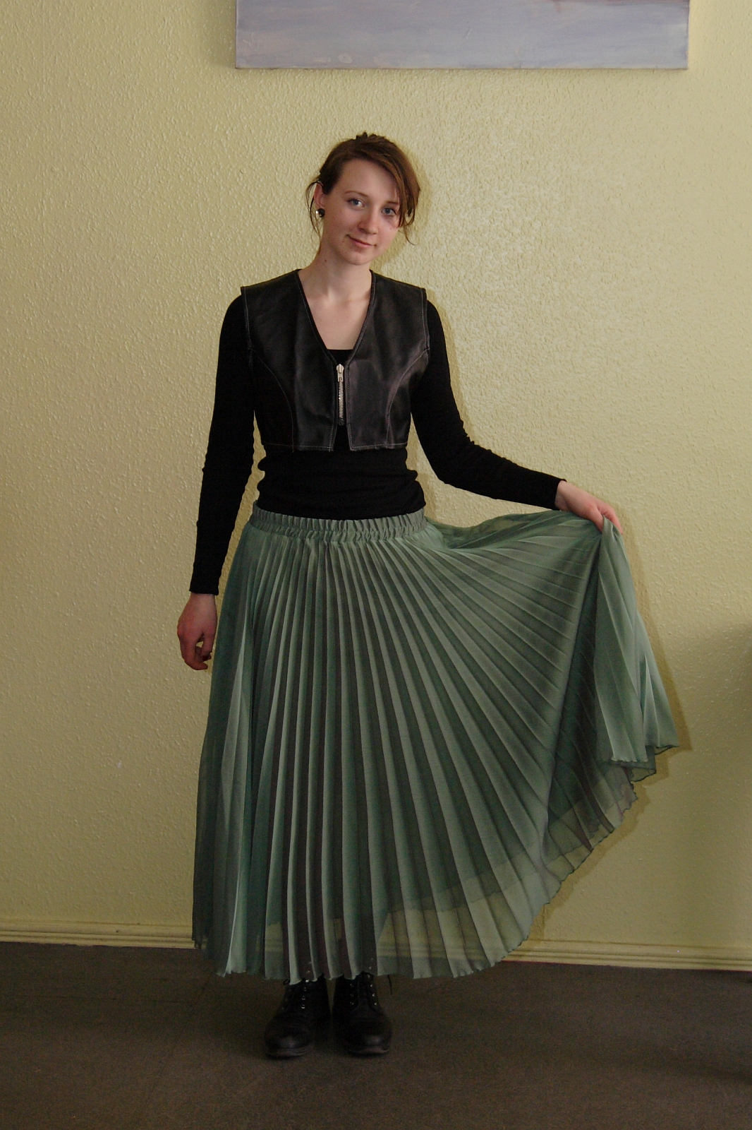 A person stands against a yellow wall, wearing a black long-sleeve shirt with a sleeveless, black leather-look vest from Talka Modes, and a flowing, pleated green skirt. They hold the skirt delicately with one hand and wear black lace-up boots. Their hair is pulled back, and they are smiling slightly.