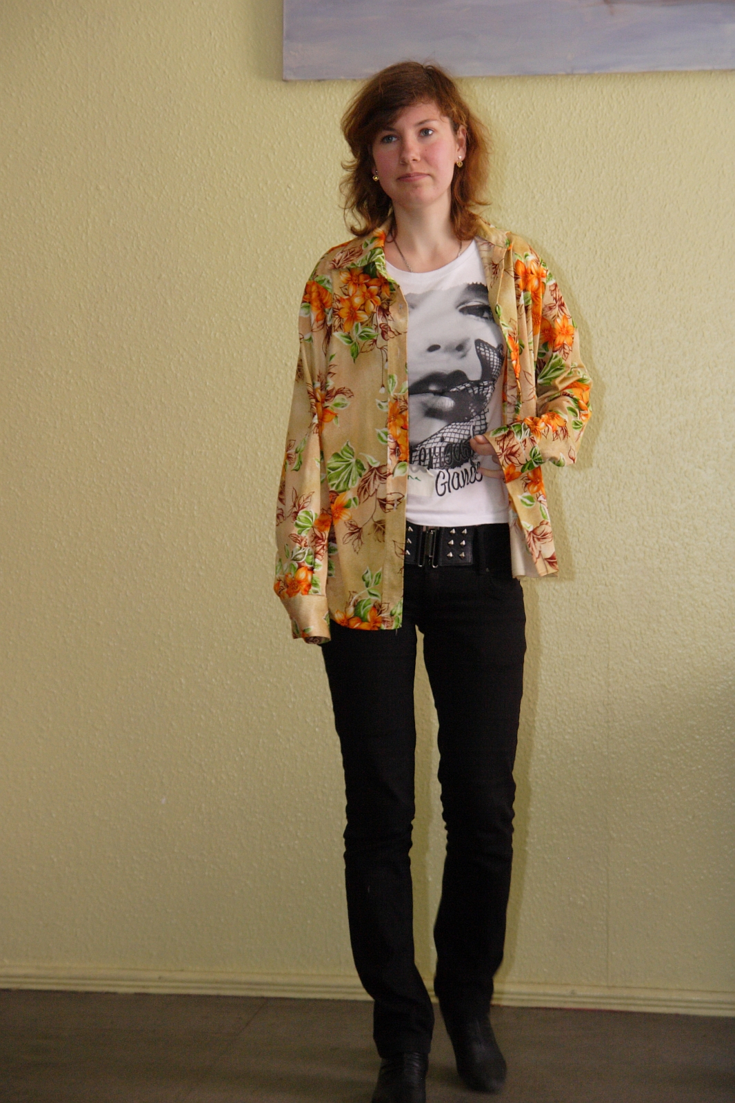 A person with shoulder-length hair is standing indoors against a light-colored wall. They are wearing a bright floral shirt over a graphic t-shirt, black pants, and black shoes. Adopting a relaxed mode, they look straight ahead with a neutral expression and have one hand partially in a pocket.