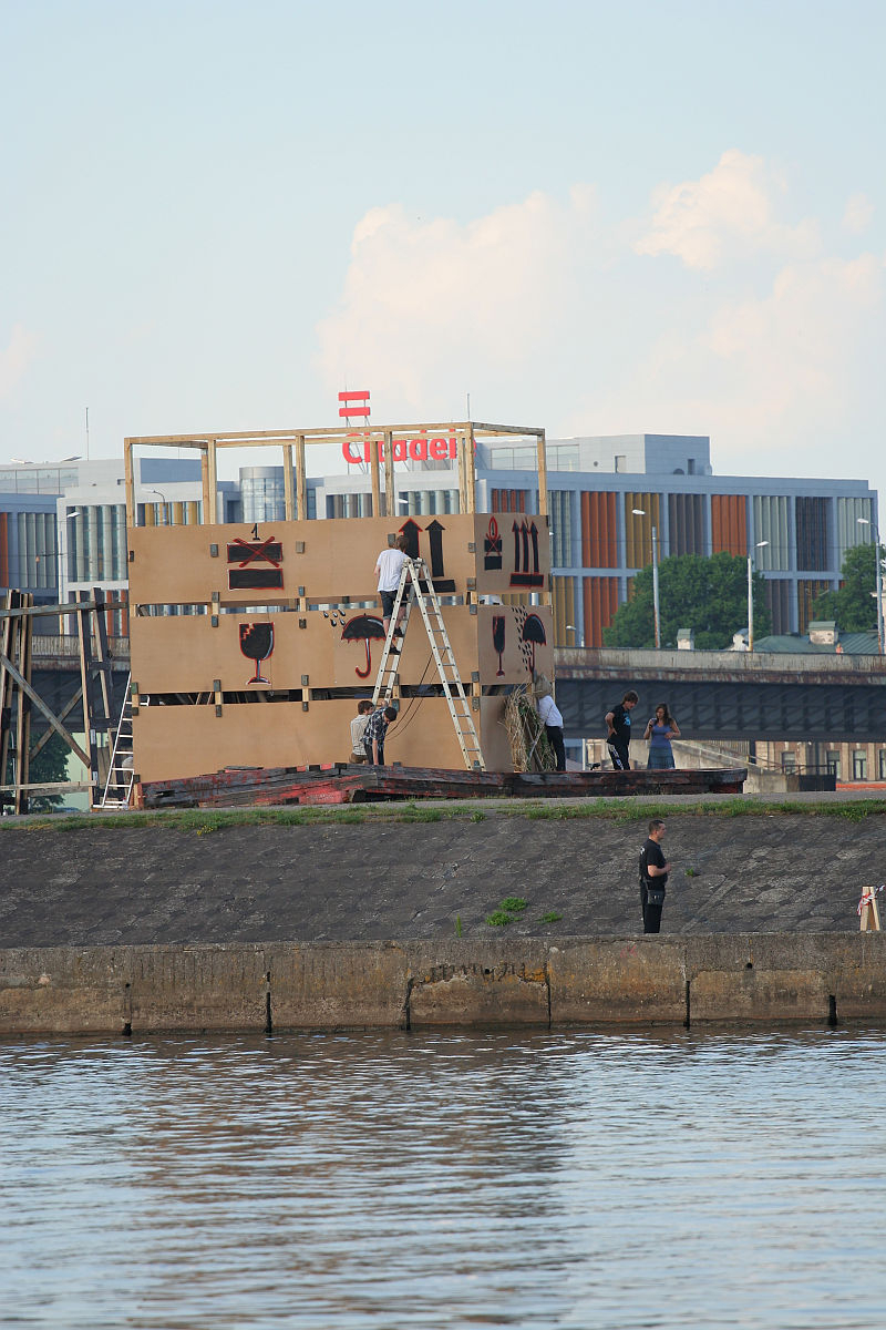 Several people are assembling a large wooden structure resembling part of a building facade along a riverside during the Kapusvētki event. Two individuals are on ladders adjusting the structure. In the background, modern buildings are visible. One person stands near the waterway, with greenery in the distance.