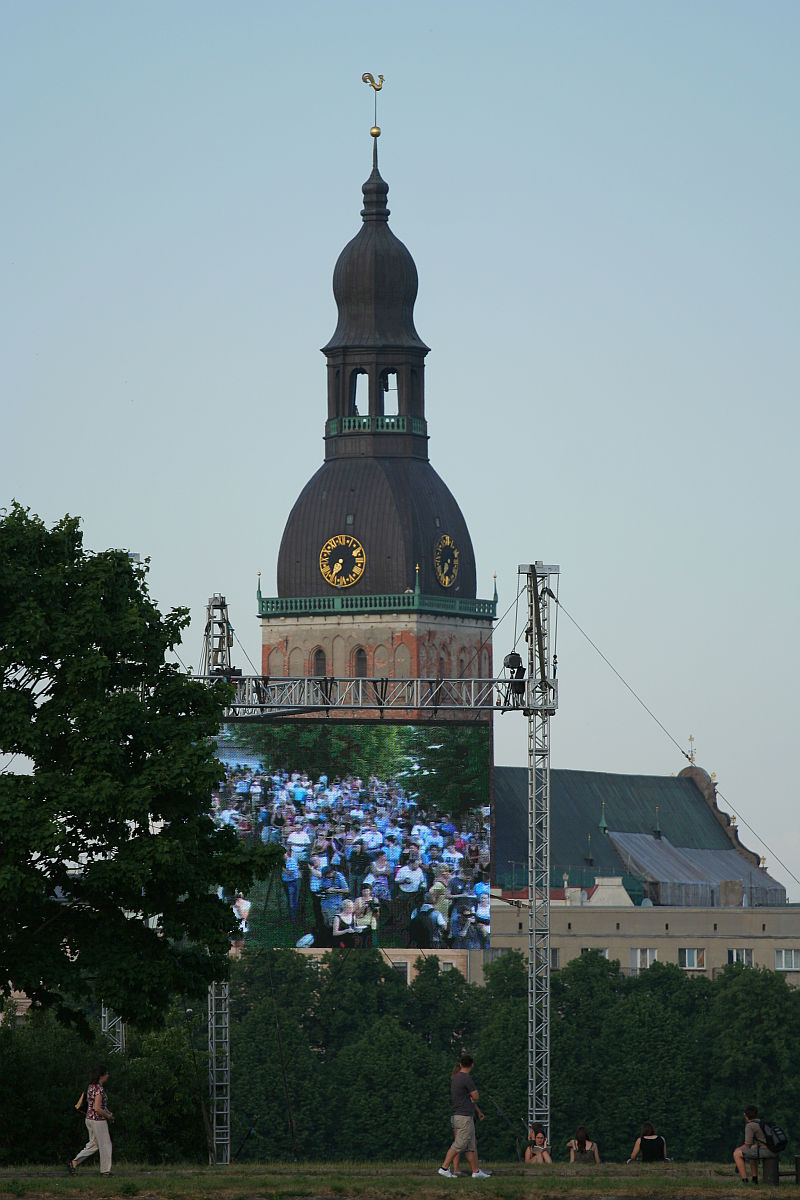 A tall, dome-topped church tower with a weather vane rises against a clear blue sky. Below, a large screen shows a crowd of people, possibly at Kapusvētki. The setup is outdoors, with tree foliage and scattered onlookers in the foreground. The scene conveys a blend of historic architecture and modern technology.