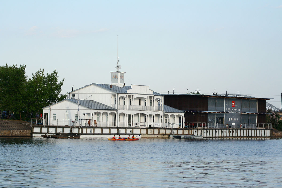 A serene waterfront scene showcases a white, multi-story boathouse with a clock tower and veranda. Beside it, a dark, modern building with "BRIGANTIA" signage stands tall. Two people in a red-orange kayak paddle on the calm water in the foreground, enjoying the tranquility of Kapusvētki. Trees flank the buildings under a clear sky.