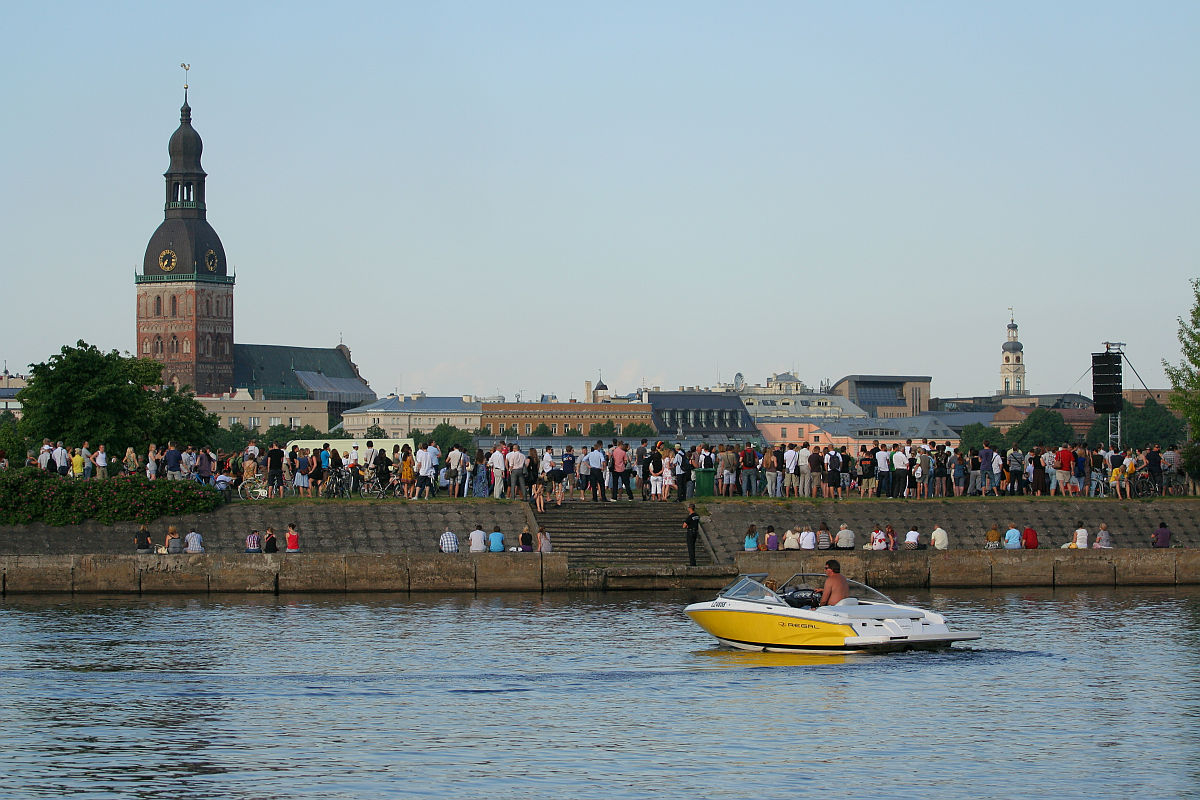 A yellow and white speedboat with a single person on board cruises along a river in the foreground. In the background, numerous people are gathered along the riverbank near a historic church with a tall clock tower, celebrating Kapusvētki. The sky is clear and light blue, indicating a calm and pleasant day.