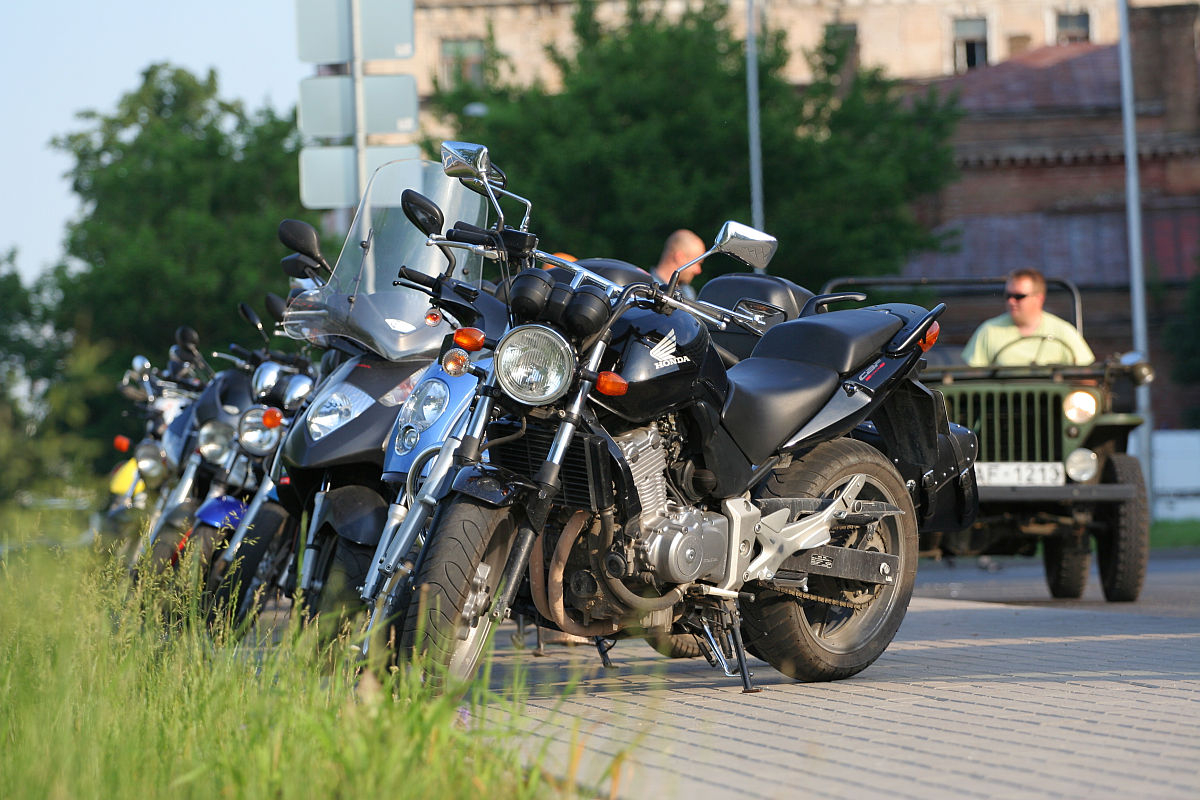 Several parked motorcycles with various designs and models, including a Honda, are lined up on the side of the road near a grassy area. In the background, an old vehicle is seen, and two people are near the motorcycles. The scene resembles a festive kapusvētki gathering with buildings and greenery visible in the distance.