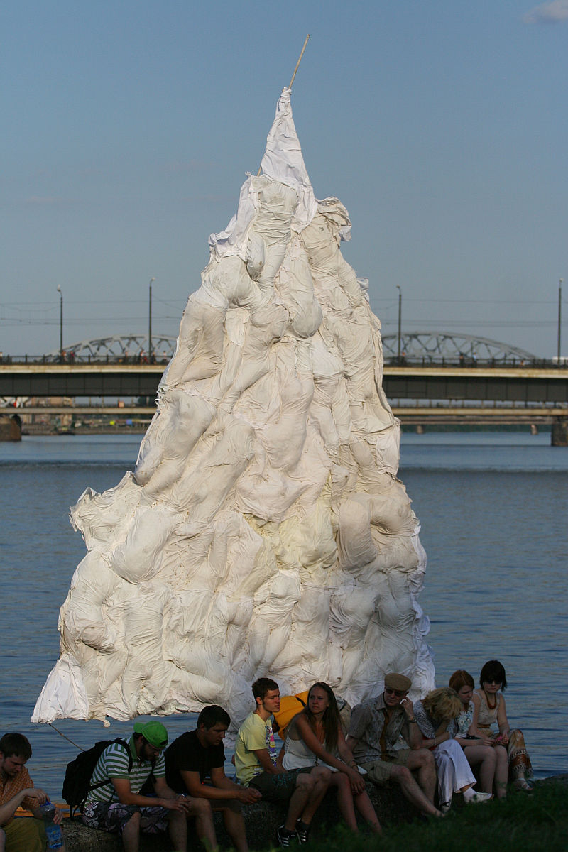 A group of people sit next to a river beneath a large, white, conical art installation made of fabric. Reminiscent of the Oligarhu kapusvētki celebration, the structure resembles a tree or mountain and is held up by a central pole. In the background, a bridge spans across the river under a clear blue sky.