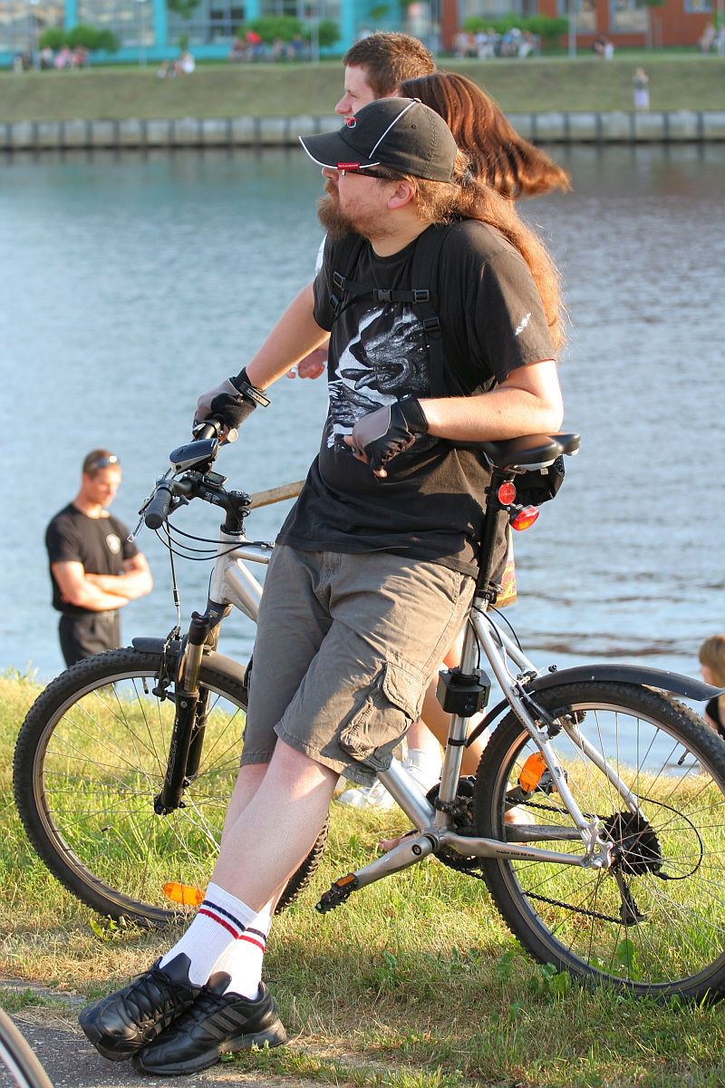 A person with long hair sits on a gray bicycle near a riverbank during kapusvētki. They wear sunglasses, a black T-shirt with a dragon design, gray shorts, gloves, and black shoes with white socks. Two other people stand nearby looking out over the water. The background includes more people and a green lawn by the water.