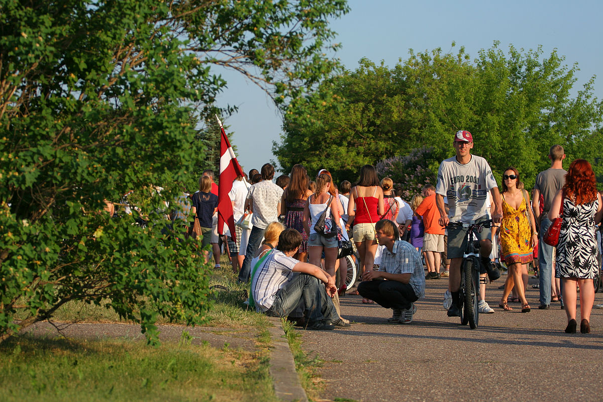 A crowd of people enjoying a sunny day in a park during the Oligarhu kapusvētki. Some are walking, while others are sitting or cycling. A young man wearing a cap is riding a bicycle, and two young men are sitting by the path. In the background, a person holds a red and white flag. Lush green trees surround the scene.