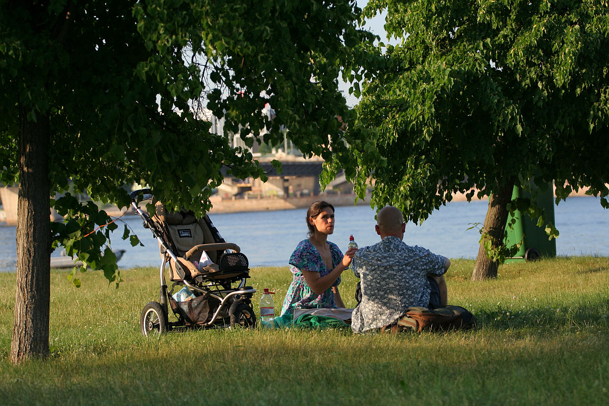 A couple is sitting on the grass under a tree, by a river. The woman in a blue top is holding a bottle and talking to the man in a patterned shirt. A stroller with a child inside is parked nearby. In the background, there are trees and a bridge over the river shining brightly like kapusvētki celebrations.