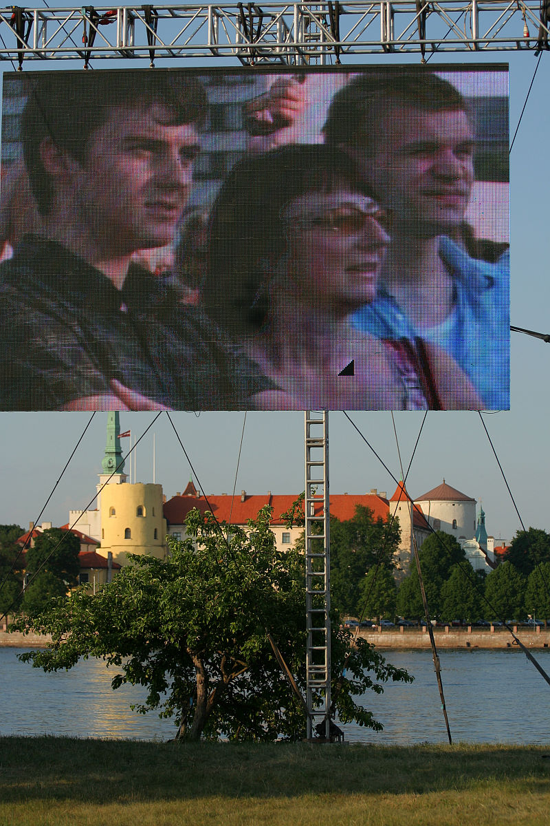A large outdoor screen displays a close-up of three people, two men and a woman, attentively watching something. The screen is mounted on a metal frame by the river during Kapusvētki, with a scenic background of historic buildings and greenery. The blue sky adds to the pleasant atmosphere.