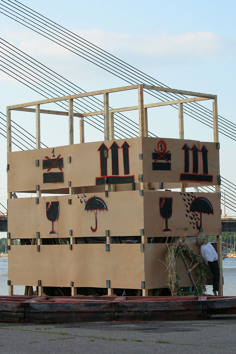 A large, unfinished wooden structure adorned with wooden panels stands by a body of water. The panels feature red and black stencil art, including designs of umbrellas, wine glasses, and musical notation. Celebrating kapusvētki nearby, in the background, a bridge with cables stretches across the sky.