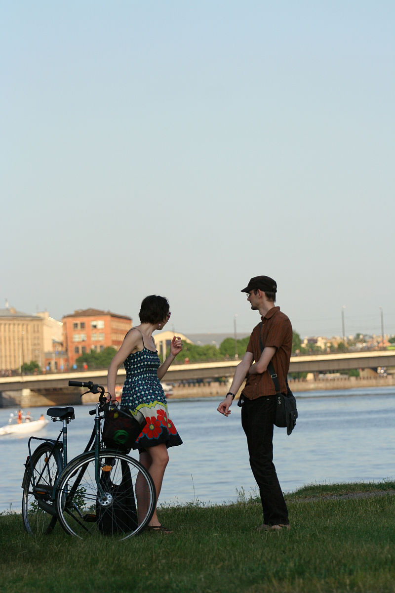 Two people are standing by a riverbank next to a bicycle, engaged in conversation. The person on the left wears a colorful dress and gesticulates while the person on the right, dressed in casual attire with a hat, looks on. Behind them, during Kapusvētki celebrations, a boat is on the water with buildings in the background.