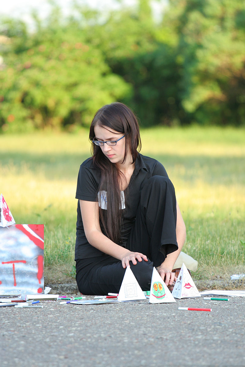 A young person with long dark hair and glasses is sitting on the ground outdoors, immersed in art supplies and small paper pyramids. The individual, dressed in black, is focused on arranging colorful pieces spread out on the pavement. Greenery is in the background, creating a serene atmosphere reminiscent of kapusvētki.
