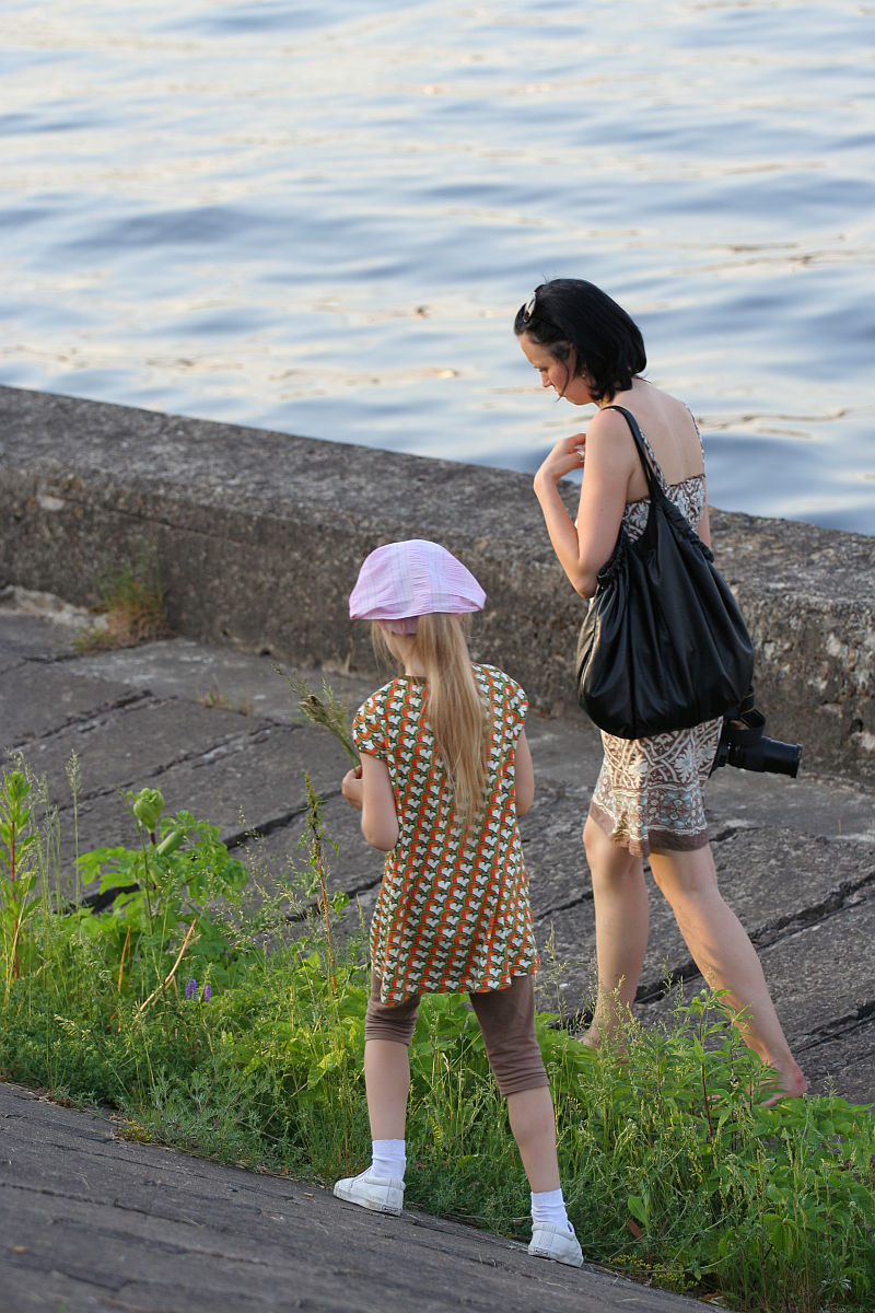 A woman with short dark hair, wearing a patterned dress and carrying a large black bag, walks along a concrete path by the water during Kapusvētki. She is accompanied by a young girl with long blonde hair, wearing a pink hat, brown leggings, and a patterned dress. The girl is looking at the plants by the path.