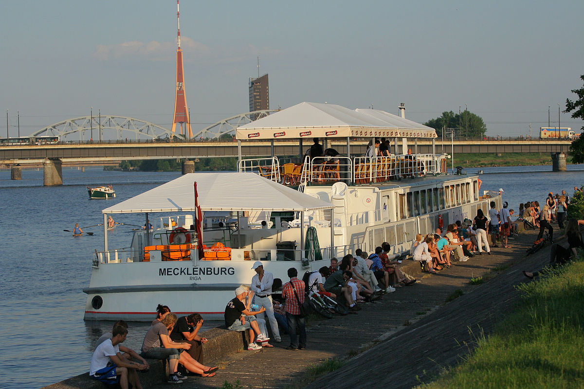 A large riverboat named Mecklenburg is docked beside a promenade on a sunny day during Kapusvētki. Many people are seated and standing along the quay, relaxing and socializing. Behind the boat, there's a bridge and a tall broadcasting tower. The scene reflects a lively, leisurely atmosphere by the river.