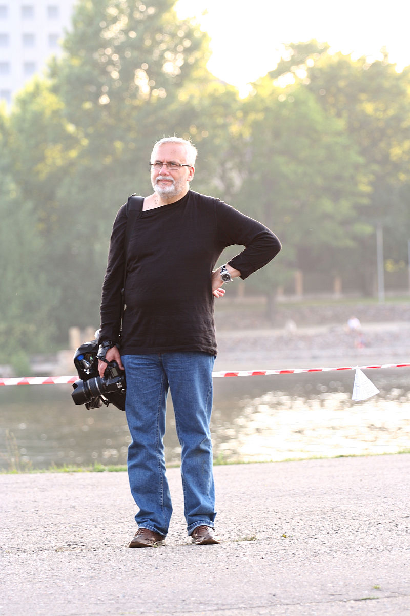 A man with gray hair and a beard stands outdoors in front of a river during Kapusvētki, wearing a black long-sleeve shirt and blue jeans. He holds a professional video camera in his right hand and has his left hand on his hip. The background includes trees, a building, and a red-and-white striped barrier.
