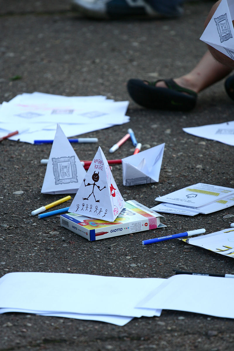 An outdoor scene shows various stationery items scattered on the ground, including colored markers, papers, and a box of Giotto pencils. There are handmade paper cones decorated with drawings and text, reminiscent of Oligarhu kapusvētki celebrations. A partial view of a person's legs wearing black shoes is in the background.