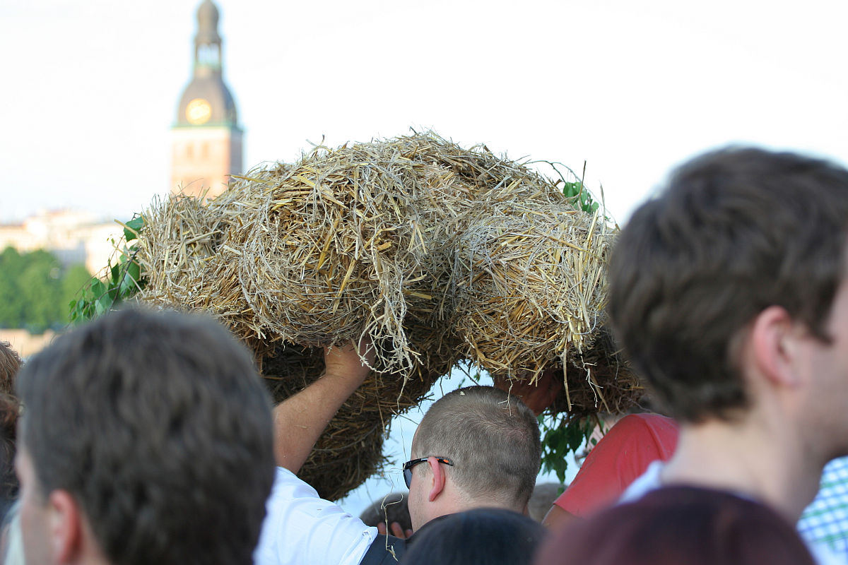 A group of people in an outdoor setting carry a large bundle of straw shaped into a figure, reminiscent of the Oligarhu kapusvētki celebration. In the background, a tall building with a clock tower is visible. The sky is clear and sunny. Some individuals have their backs to the camera, their faces not clearly visible.