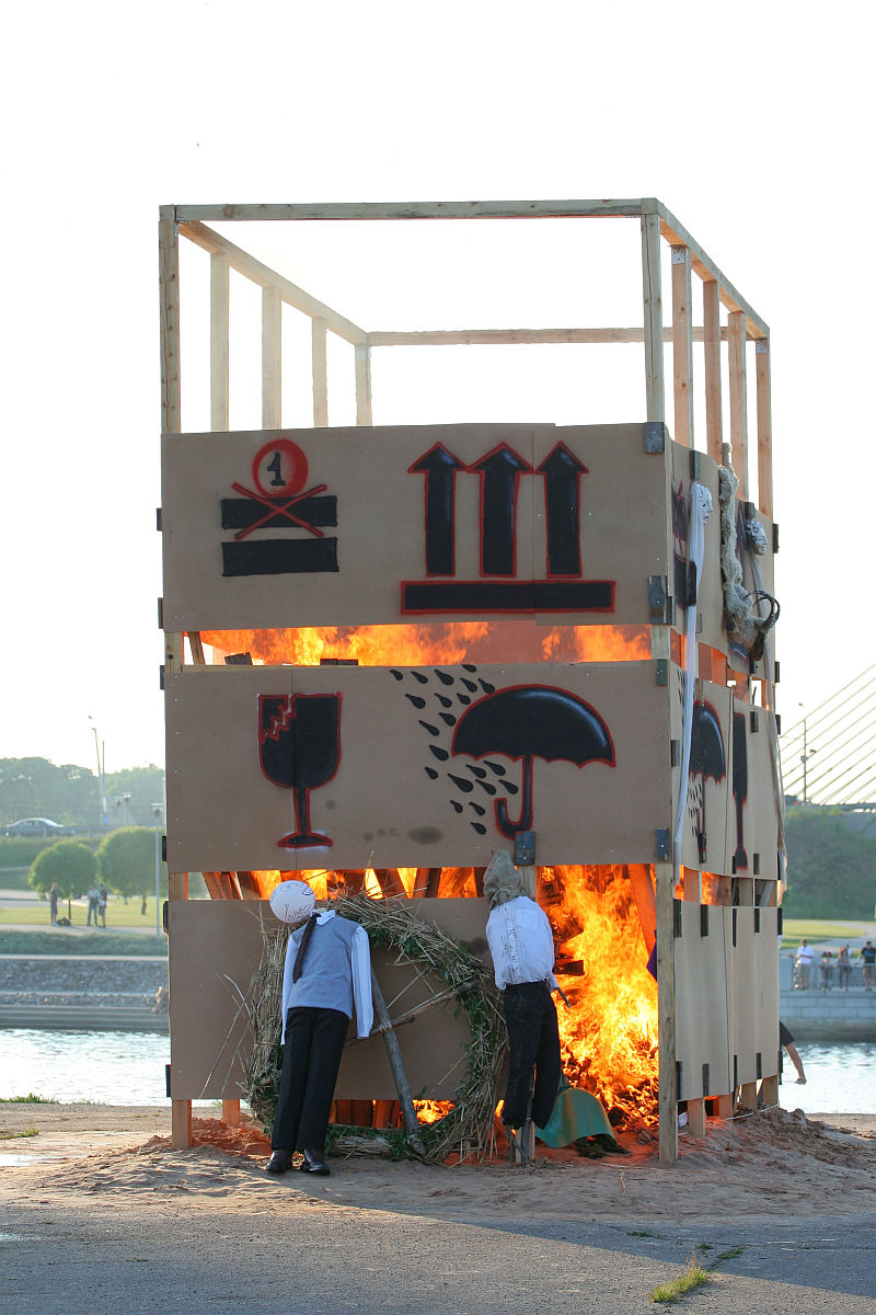 A large, rectangular wooden structure is set on fire during the Kapusvētki celebration. The structure is covered in brown panels featuring red and black painted symbols. Effigies resembling human figures wearing clothes are placed around the structure. Trees and a river with a bridge are visible in the background.
