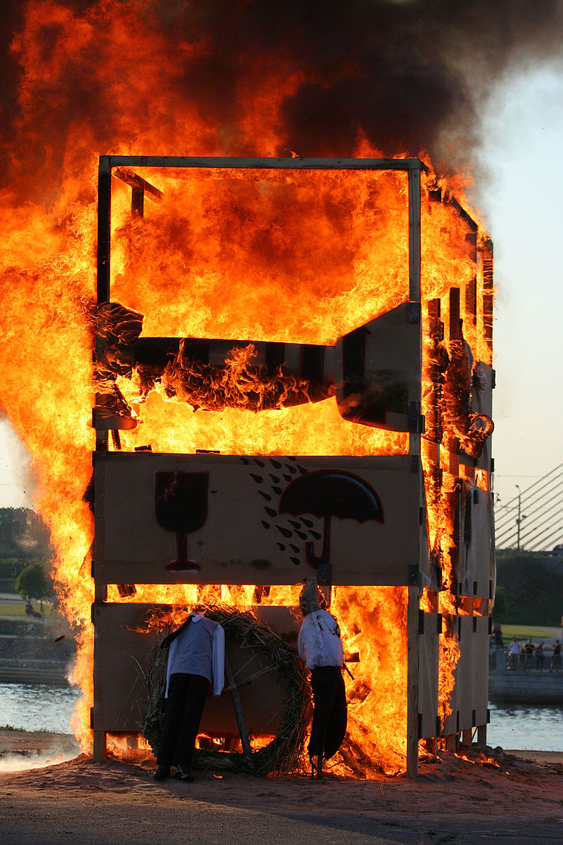 A large wooden structure is engulfed in flames, emitting thick black smoke. Two figures in white shirts and dark pants are seen near the base of the structure, one bending down and the other standing. The scene appears to be part of kapusvētki festivities by a riverside setting with a bridge in the background.