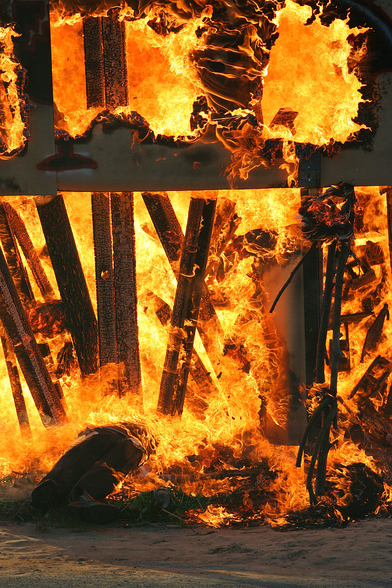 A two-story structure engulfed in intense flames during what appears to be a controlled burn or fire training exercise for the upcoming Kapusvētki festival. The fire consumes wood beams and debris, creating thick, bright orange flames and plumes of smoke. The scene is chaotic with the fire spreading rapidly, engulfing the entire structure.