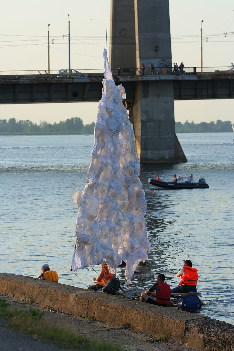 People are gathered near a large, abstract installation resembling a tree made of white fabric. Set beside a calm river during the Kapusvētki festival, a bridge and some pedestrians form the backdrop. A small motorboat, possibly belonging to an Oligarhu, is visible on the water near the installation.