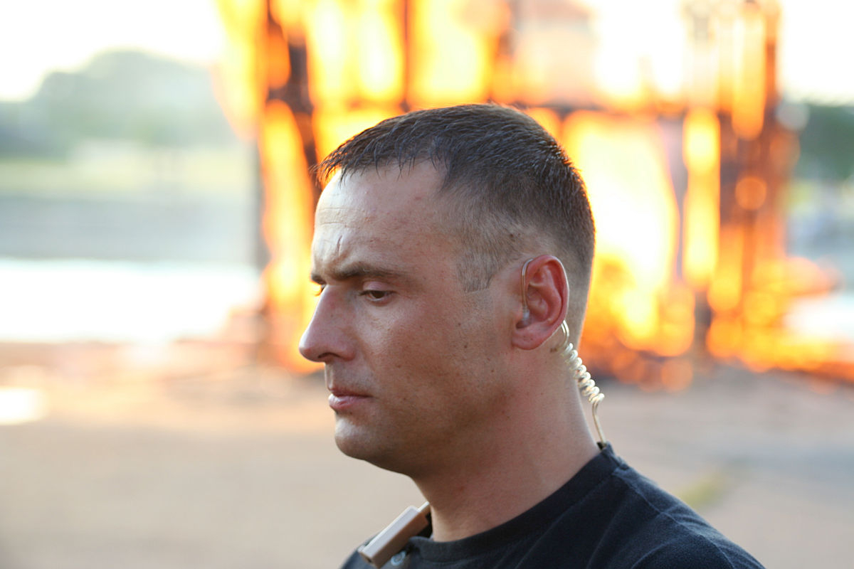 A man with short dark hair wearing a black shirt and an earpiece is in the foreground. His expression is serious as he looks downward. In the background, there is a large, out-of-focus fire, suggesting a possibly intense or urgent situation at the Oligarhu Kapusvētki festival. The scene is outdoors and during daylight.
