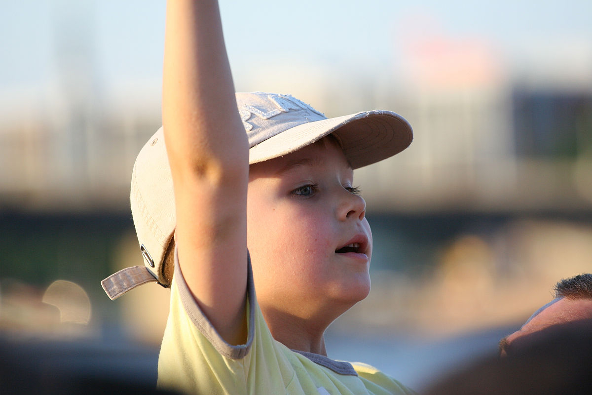 A young child wearing a light-colored baseball cap and a yellow shirt is shown raising their right arm at the Oligarhu kapusvētki. The child looks engaged or excited, with mouth slightly open, looking towards the right. The background is blurred, with indistinct structures and a blue sky.