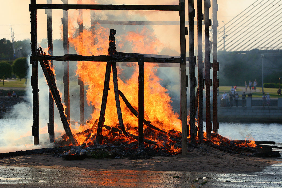 A large wooden structure engulfed in flames by a waterfront during the Oligarhu kapusvētki. The tall, burning wooden beams are partially collapsed. Smoke rises into the sky as people on the other side of the water observe. A modern bridge with cables is also visible in the background.