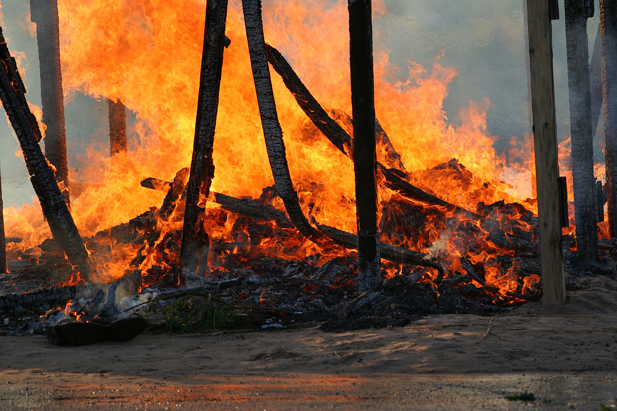 A scene of intense fire consumes wooden structures during kapusvētki. Bright orange and yellow flames engulf charred beams as smoke billows into the dirt-laden air. The ground, covered in ash and debris, tells a somber tale of destruction in the shadow of Oligarhu.