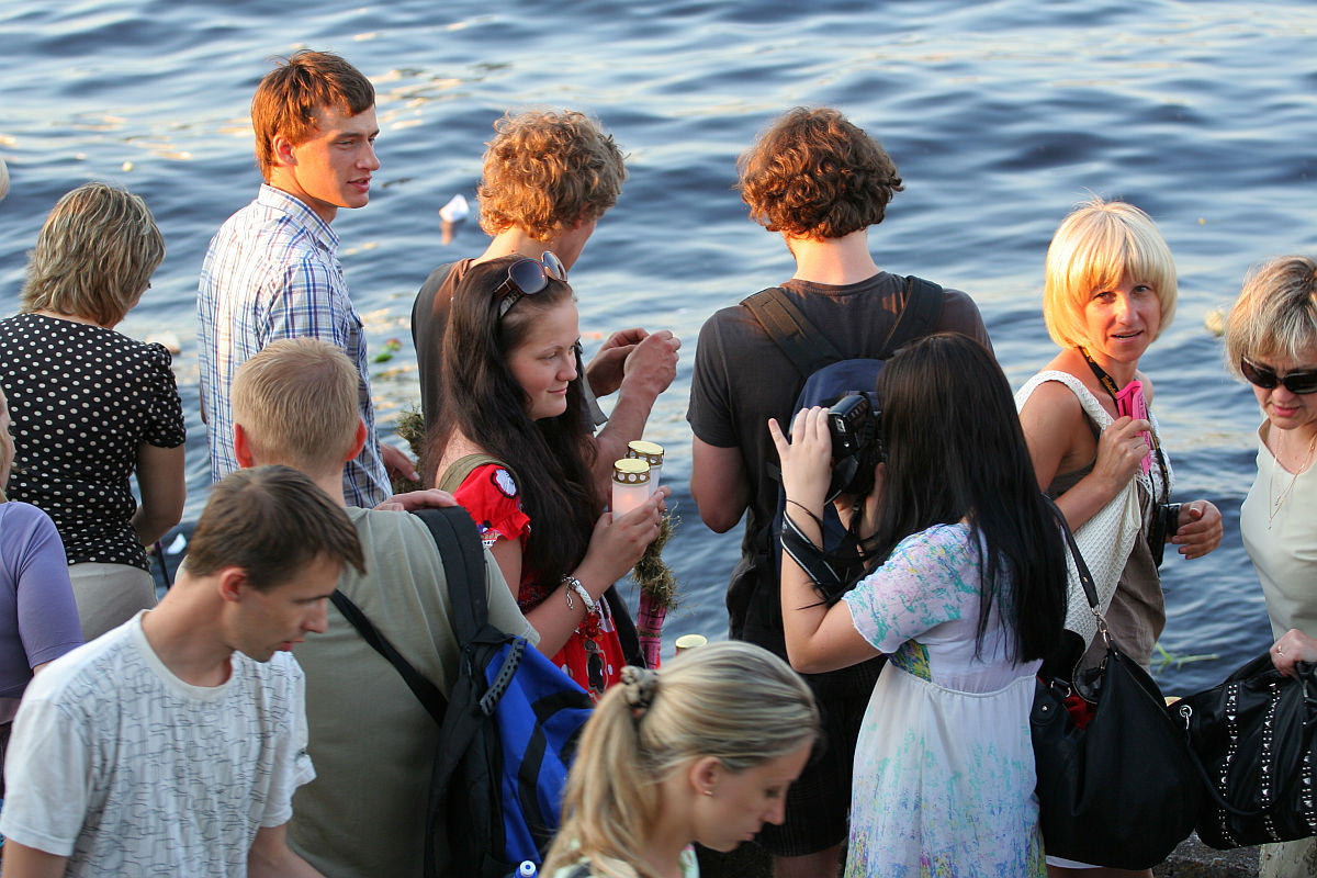 A group of people, including young men and women, are gathered near a body of water during kapusvētki. Some are taking photos, others are engaged in conversation, and a few are holding ice cream cones. They are casually dressed, and the water in the background suggests a relaxed, outdoor setting.