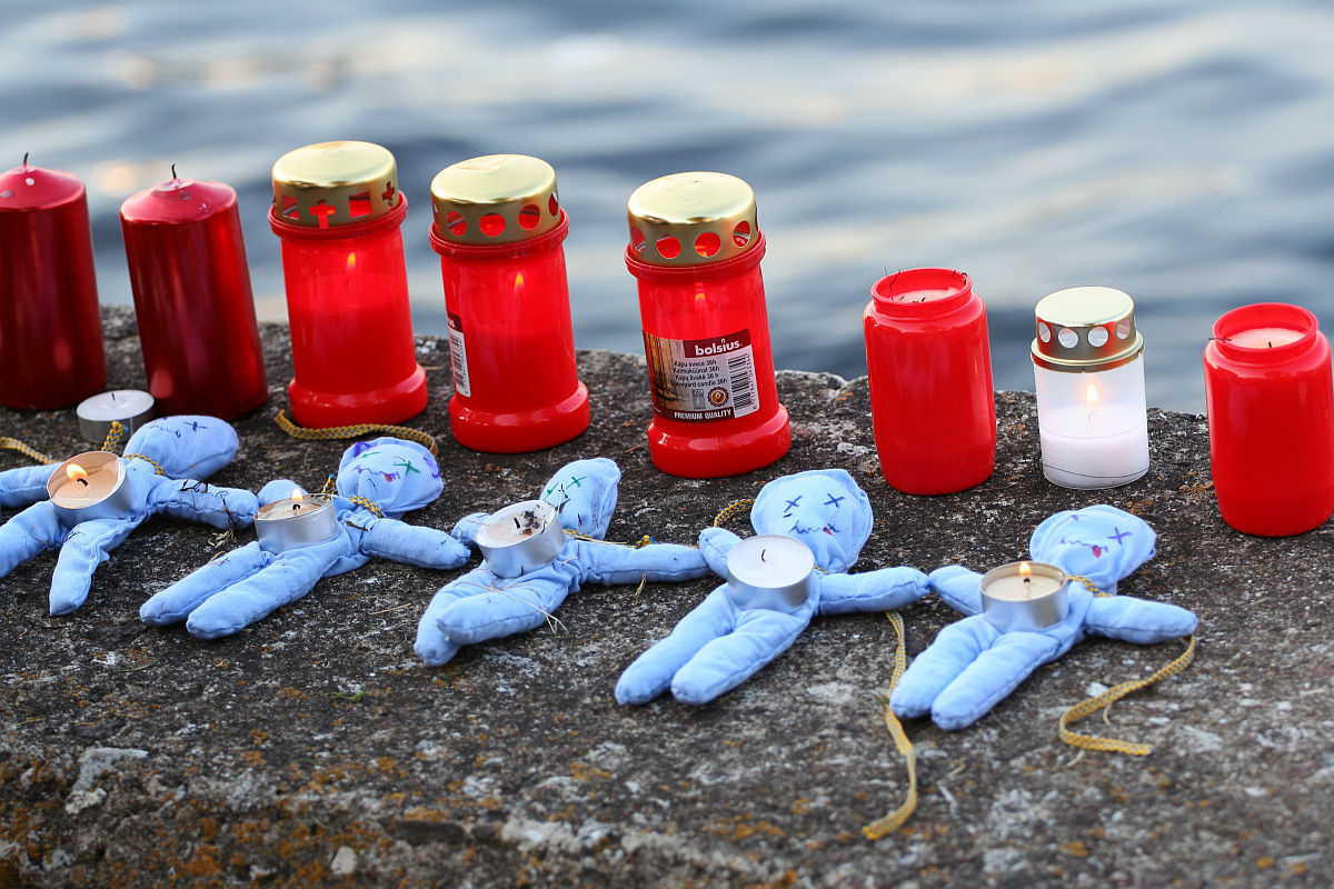 Several red candle jars and lit tealights are arranged in a row on a stone ledge by the water. Four blue fabric dolls with tealights placed on their midsections are positioned in front of the candles, suggesting an Oligarhu or kapusvētki tribute. The scene appears to be a memorial setup.